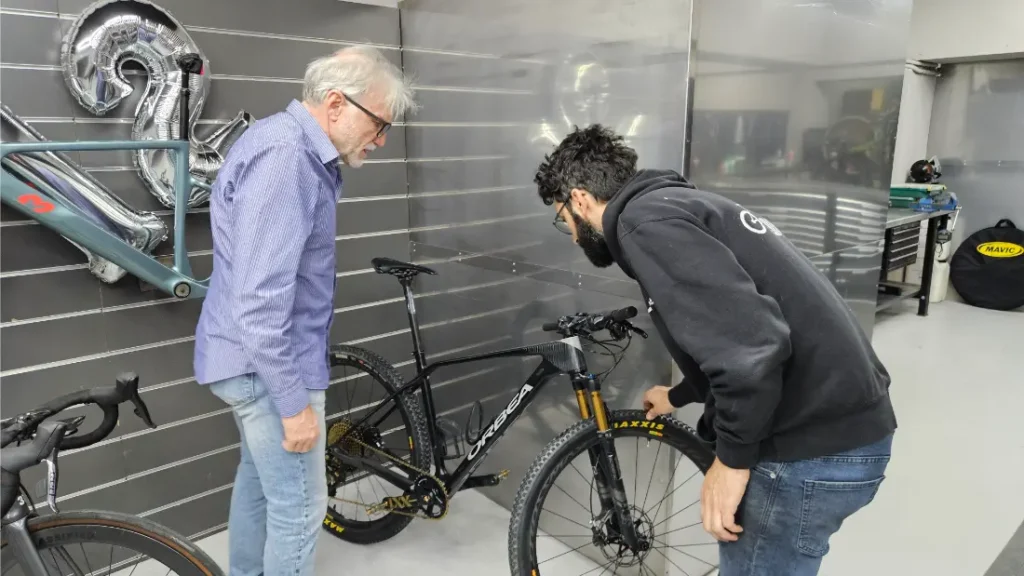 Dos hombres examinan una bicicleta de montaña negra en un taller de Biciclinic con paredes grises, un cuadro de bicicleta en la pared y globos formando el número 29 en el fondo.