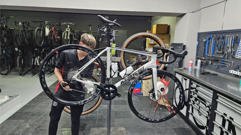 Una persona realiza mantenimiento a una bicicleta de carretera blanca montada en un soporte de reparación en el taller de Biciclinic, con herramientas y otras bicicletas al fondo.