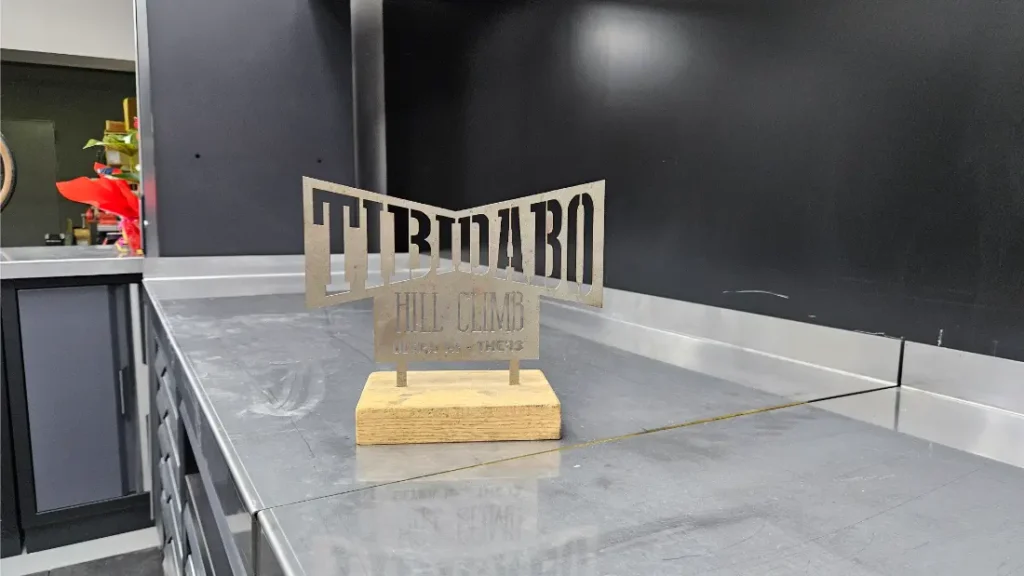 En el entorno industrial del taller de Biciclinic se exhibe un cartel metálico “SUBASTA AL TIBIDABO” con base de madera sobre un banco de trabajo de acero inoxidable.
