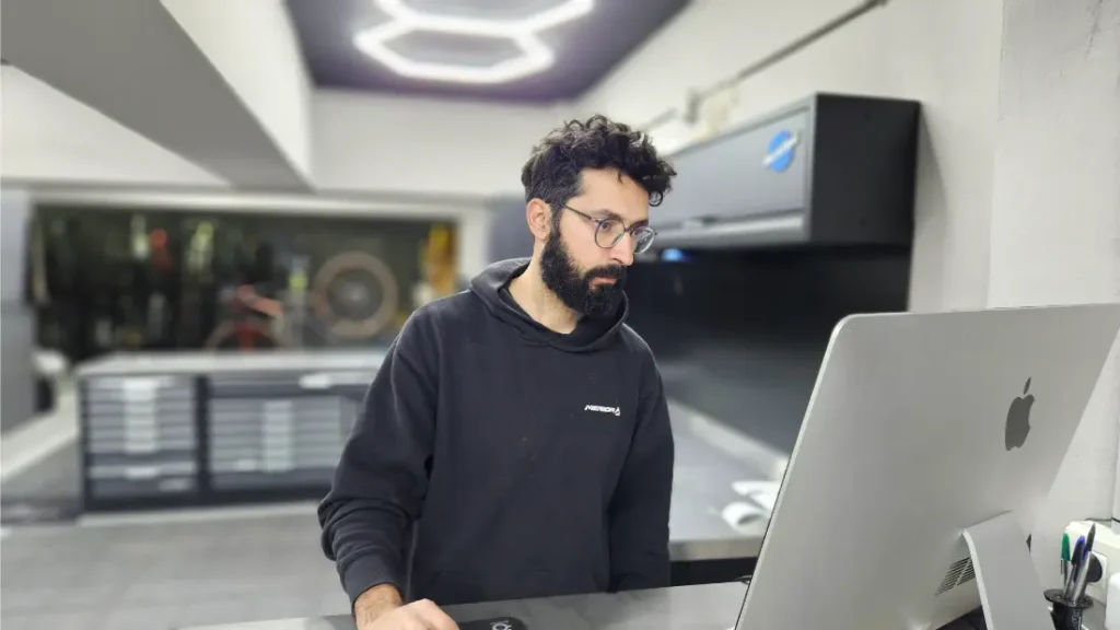 Un hombre con cabello oscuro y barba, que usa anteojos y una sudadera con capucha negra, está de pie frente a un escritorio usando una computadora Apple grande en un espacio de trabajo moderno y bien iluminado.