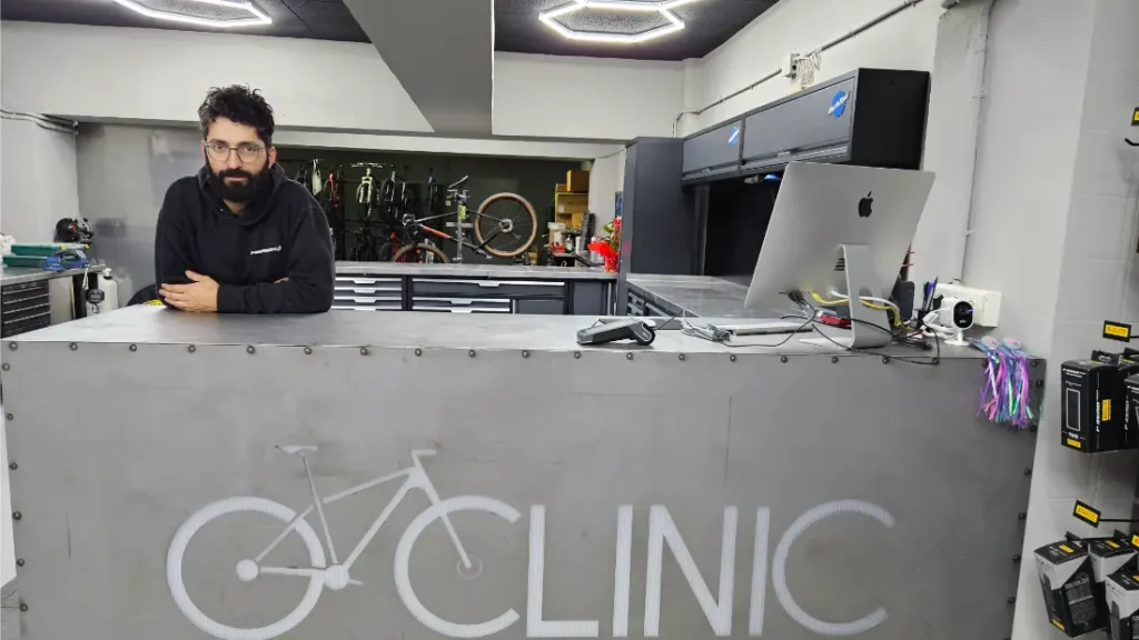 Un hombre está detrás de un mostrador metálico con el gráfico de una bicicleta y la palabra "CLINIC" en Biciclinic, un taller de reparación de bicicletas. Al fondo se ven herramientas, bicicletas y un ordenador.