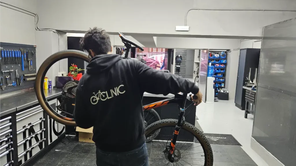 Una persona que viste una sudadera con capucha que dice "CYCLINIC" repara una bicicleta de montaña en un taller de Biciclinic bien organizado, rodeado de herramientas en las paredes y armarios de almacenamiento.