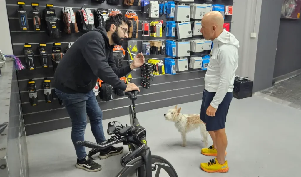 Dos hombres conversan en la tienda de bicicletas Biciclinic; uno sostiene una bicicleta y hace gestos, mientras que el otro permanece con un perro a su lado, frente a una pared de accesorios de ciclismo.