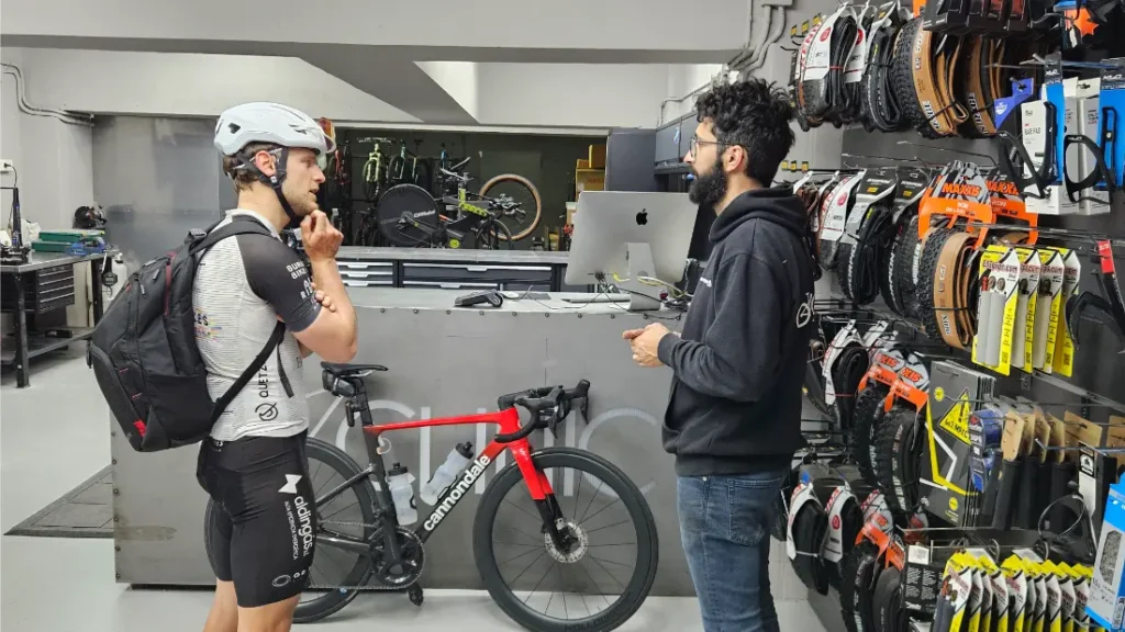 Un ciclista vestido con equipo y casco conversa con un miembro del personal en el mostrador de Biciclinic, mientras en la pared se exhiben neumáticos y accesorios de bicicleta.