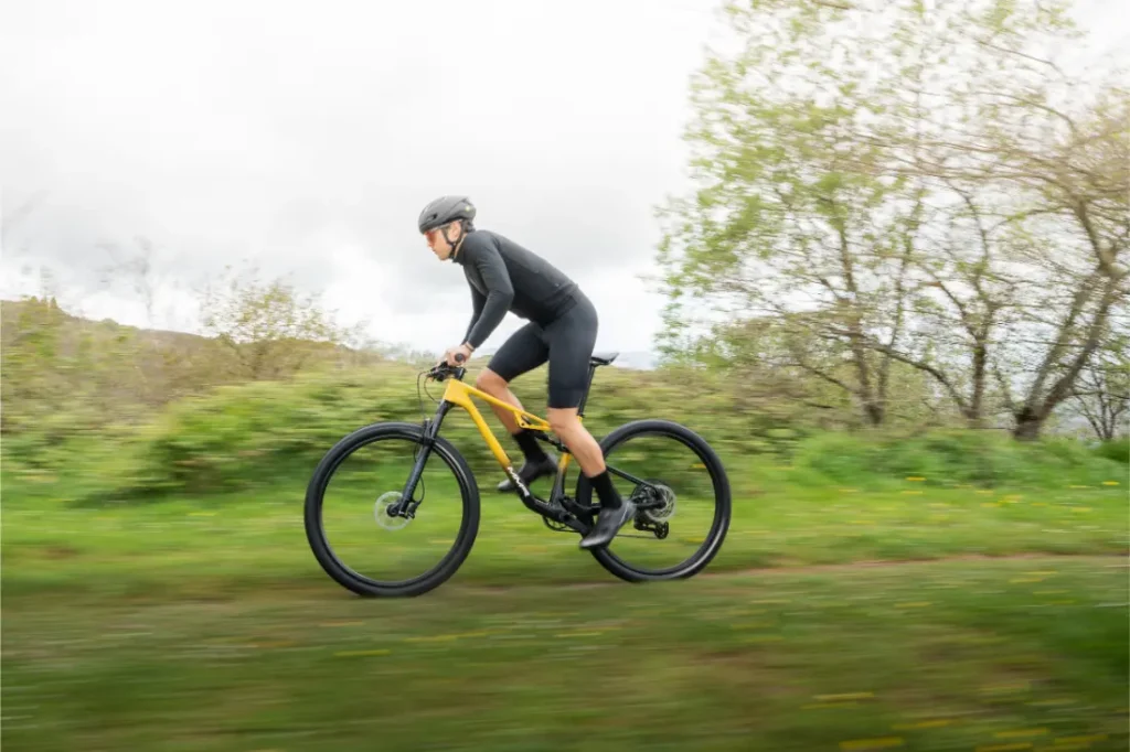 Una persona con casco y equipo de ciclismo monta una bicicleta de montaña amarilla MMR Kenta en un sendero cubierto de hierba, mostrando la evolución total, con árboles y un cielo nublado en el fondo.