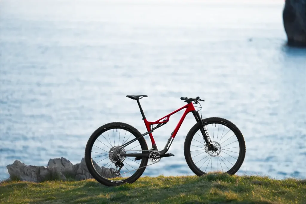 Una bicicleta de montaña roja y negra MMR Kenta, símbolo de evolución total, se encuentra sobre el césped con un cuerpo de agua al fondo.