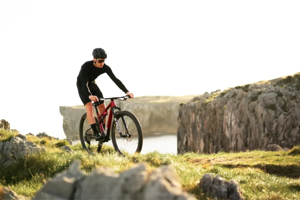 Una persona con equipo de ciclismo y casco conduce una bicicleta de montaña MMR Kenta, mostrando la evolución total en un terreno cubierto de hierba y rocoso cerca de un acantilado con agua y más acantilados detrás.