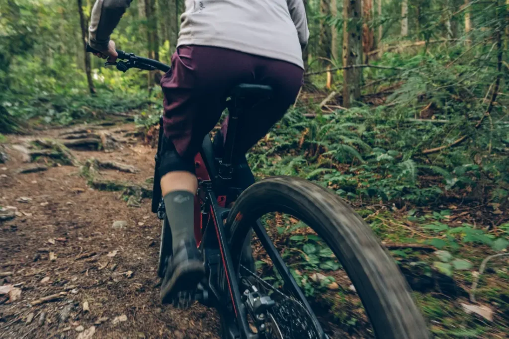 Una persona que viste una camiseta gris y pantalones cortos morados conduce una bicicleta de montaña con RockShox Reverb AXS por un sendero forestal rodeado de vegetación.