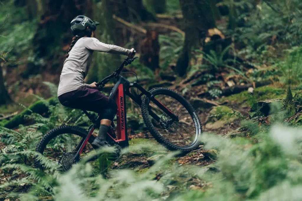 Una persona empuja su MTB cuesta arriba a través de un sendero forestal, rodeado de árboles y helechos, con el avanzado Tija RockShox Reverb AXS mejorando cada recorrido.