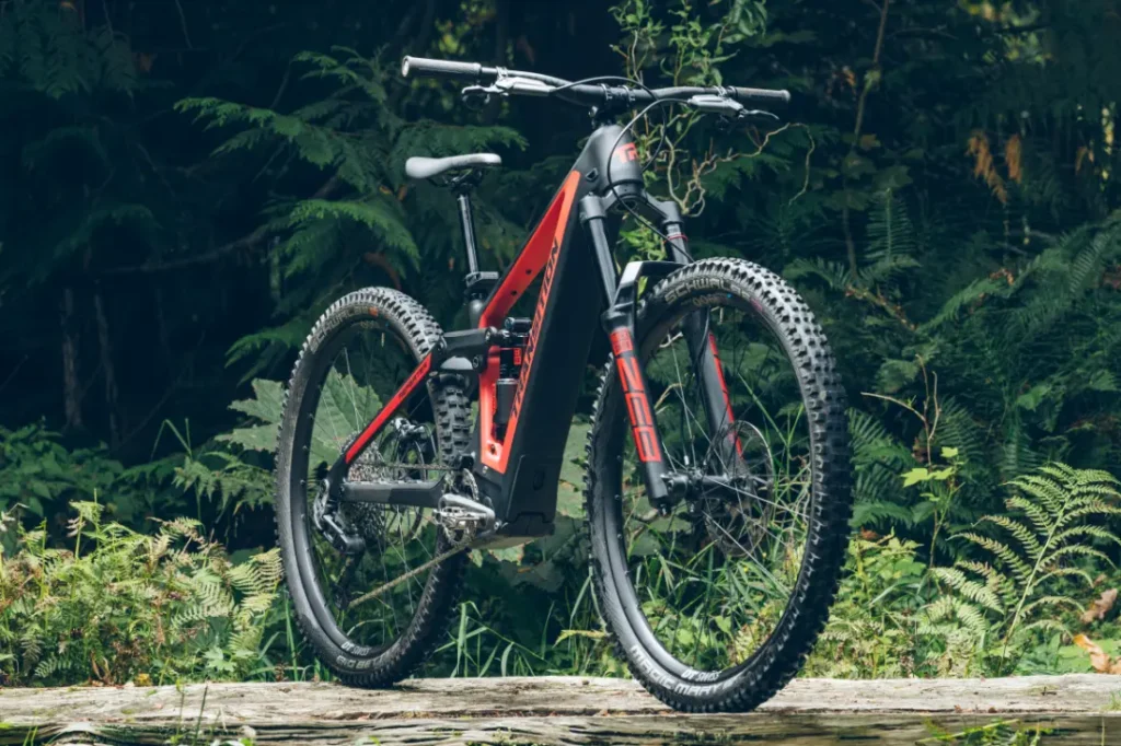 Una bicicleta de montaña negra y roja con asiento telescópico RockShox Reverb AXS está estacionada sobre una superficie de madera en una zona boscosa, rodeada de exuberantes helechos y árboles imponentes.