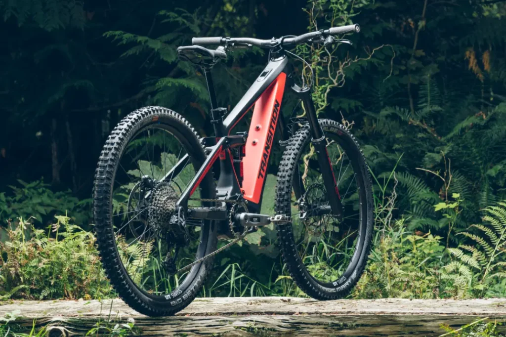Una MTB roja y negra con neumáticos gruesos se encuentra sobre una superficie de madera en un entorno forestal, equipada con RockShox Reverb AXS para un control de conducción perfecto.