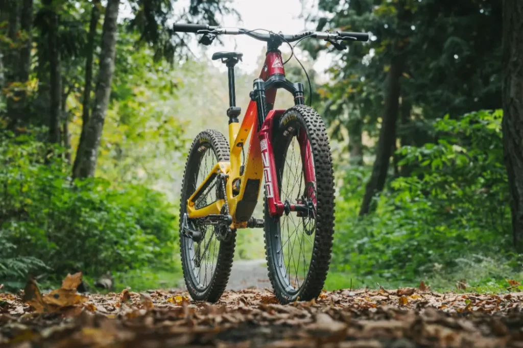 Una MTB roja y amarilla con RockShox Reverb AXS se encuentra en un sendero forestal cubierto de hojas y rodeado de árboles altos.