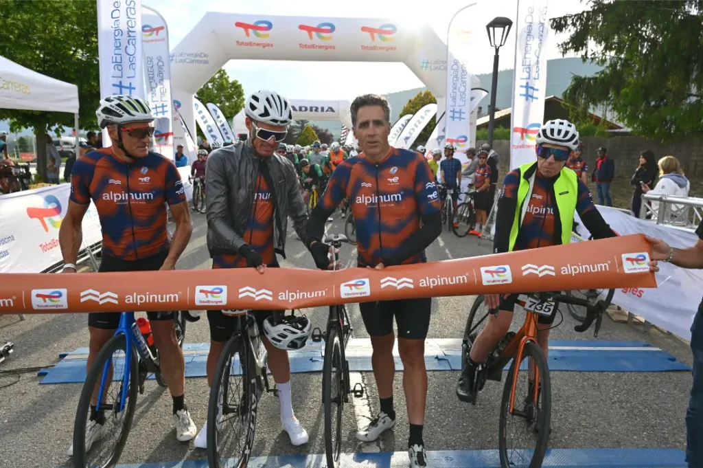 Cuatro ciclistas con camisetas Alpinum TotalEnergies de color naranja y azul a juego se encuentran con sus bicicletas en la línea de partida, preparándose para una carrera.