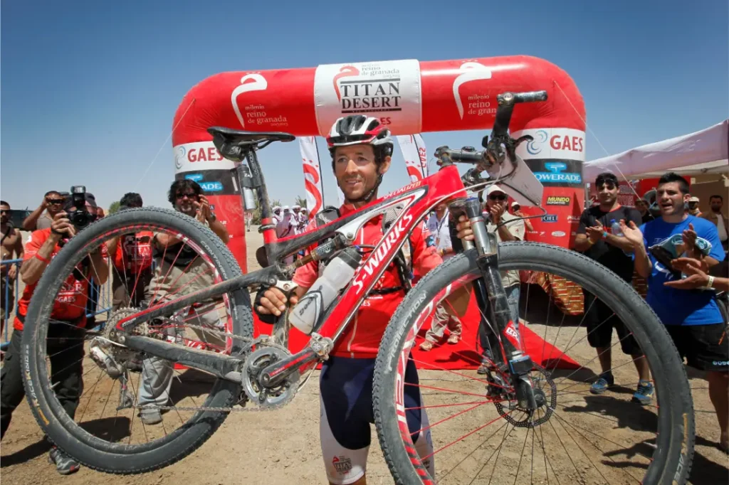 Un ciclista con ropa de carrera sostiene una bicicleta de montaña frente al arco inflable rojo Alpinum TotalEnergies en la línea de meta, rodeado de gente que lo anima y fotógrafos.