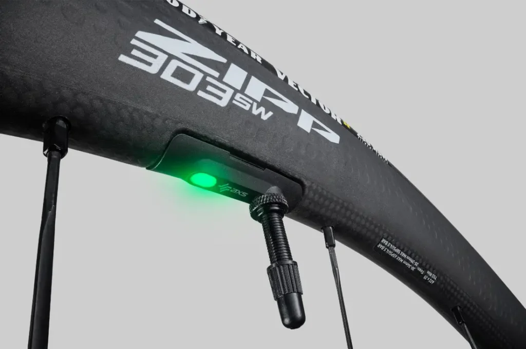 Primer plano de una rueda de bicicleta Zipp 303 de fibra de carbono con una válvula de neumático y un dispositivo que emite una luz verde, que ofrece conectividad inteligente para ayudarte a rodar más rápido.