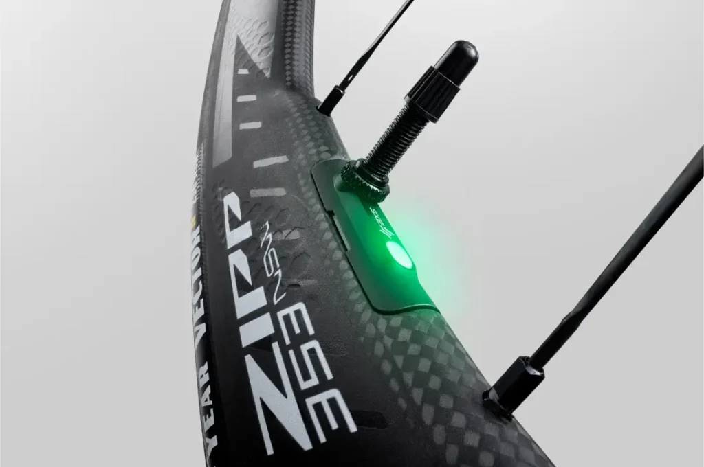 Primer plano de una rueda de bicicleta de carbono Zipp 2026 con un dispositivo sensor iluminado en verde adjunto, que muestra conectividad inteligente y dos radios visibles, diseñados para ayudarte a rodar más rápido.