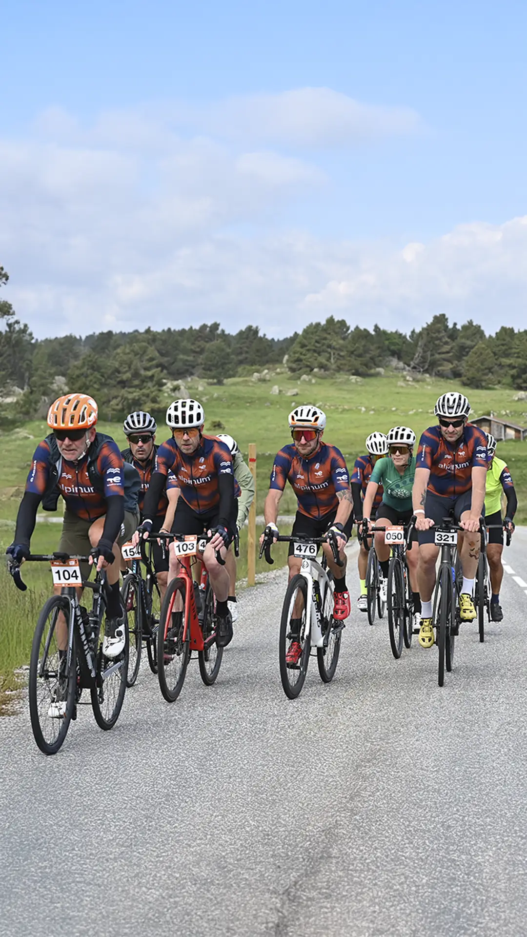 Un grupo de ciclistas que visten camisetas y cascos iguales pedalean juntos por una carretera pavimentada a través de un paisaje rural verde.