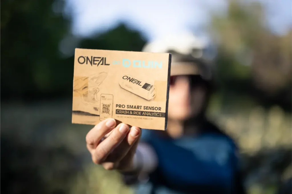 Una persona al aire libre sostiene un casco O’Neal Trailfinder Evo y una caja con sensor inteligente Quin Pro, con el producto sistema Quin y la marca MTB claramente visibles.