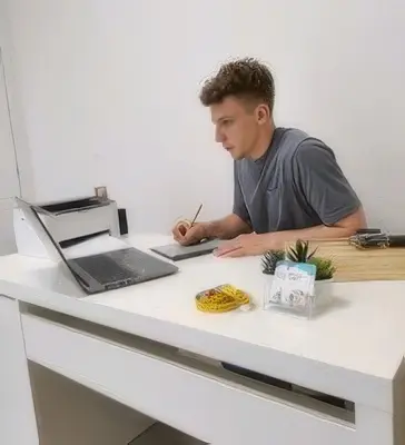 Un hombre está sentado en un escritorio blanco, escribiendo en un cuaderno mientras mira una laptop. Sobre el escritorio hay útiles de oficina, una pequeña planta y una caja; quizá tomando notas del nutricionista Bahrain Victorious, Giacomo Tomio.