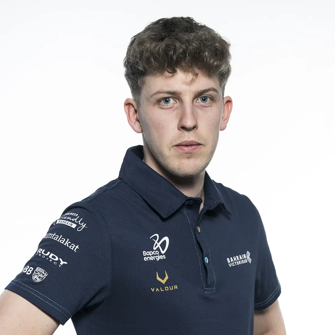 Un hombre joven con cabello corto y rizado que viste un polo azul marino con los logotipos de varios patrocinadores, incluido Nutricionista Bahrain Victorious, está parado sobre un fondo blanco liso.
