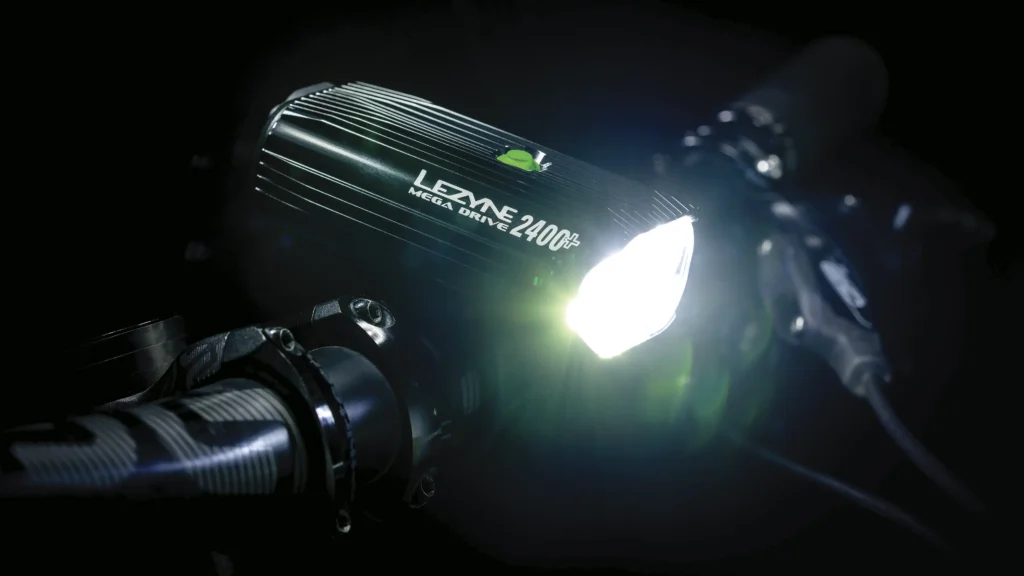 Primer plano de un faro de bicicleta Lezyne Mega Drive 2400i encendido, montado en un manillar en un entorno oscuro: algo esencial para cualquier entusiasta del Team Bike.