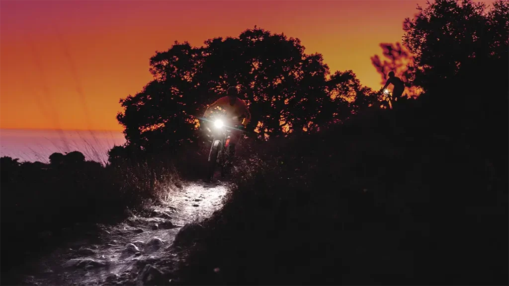 Un ciclista con una potente linterna frontal Lezyne recorre un sendero rocoso al anochecer, con la silueta de los árboles enmarcando el cielo anaranjado del atardecer. Otra persona está de pie a la derecha, miembro del equipo distribuidor de bicicletas Lezyne.