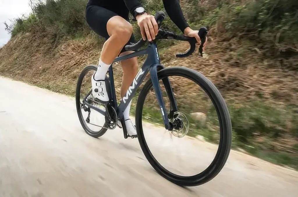 Un ciclista vestido de negro conduce una bicicleta de ruta azul MMR Simun por un camino pavimentado, rodeado de tierra y vegetación.