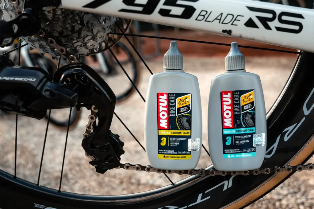 Dos botellas de lubricante de cadena Motul Bike Care y Bicimax están colocadas en posición vertical en el suelo junto a la rueda trasera y el descarrilador de una bicicleta.