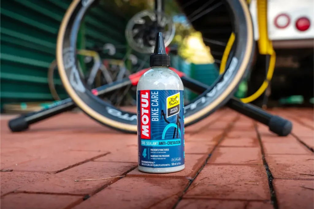 Una botella de sellador para bicicletas Motul, de la gama Motul Bike Care, reposa sobre una superficie de ladrillo, con una rueda de bicicleta y otra bicicleta Bicimax difuminadas en el fondo.