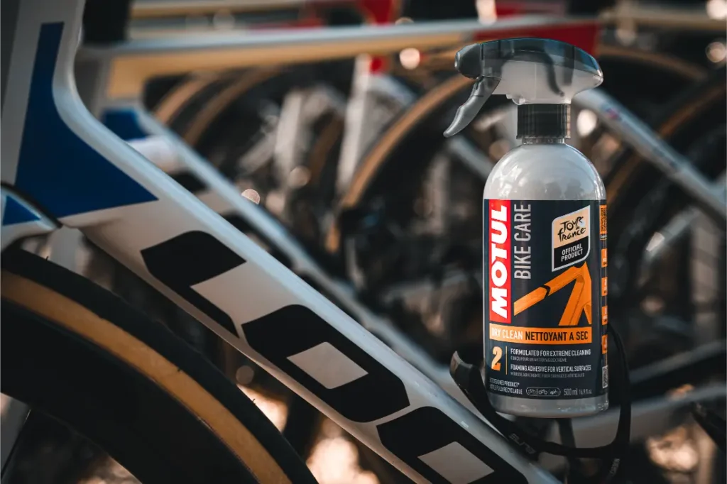 Una botella de spray limpiador Motul Bike Care se coloca contra el cuadro de una bicicleta de carretera, con varias bicicletas en el fondo, mostrando la efectiva limpieza Bicimax para los entusiastas del ciclismo.