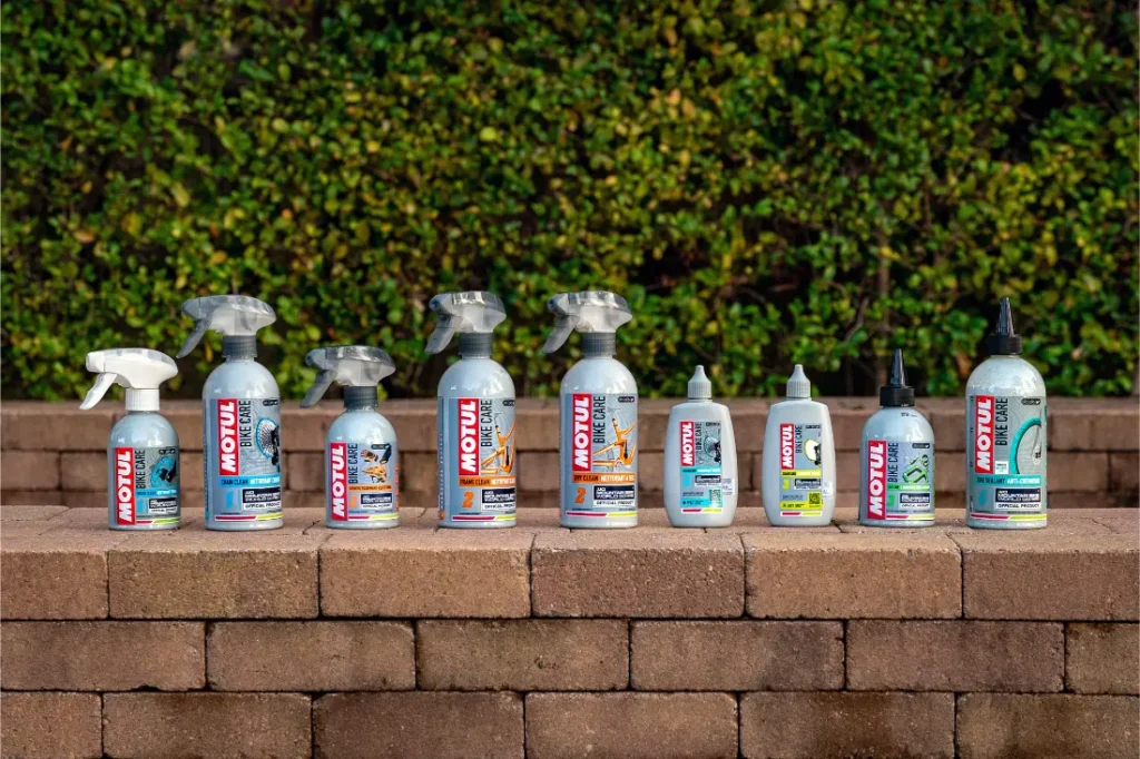 Nueve botellas rociadas y exprimibles de productos de limpieza y mantenimiento de vehículos Motul Bike Care y Bicimax están alineadas sobre una superficie de ladrillo al aire libre, con vegetación en el fondo.