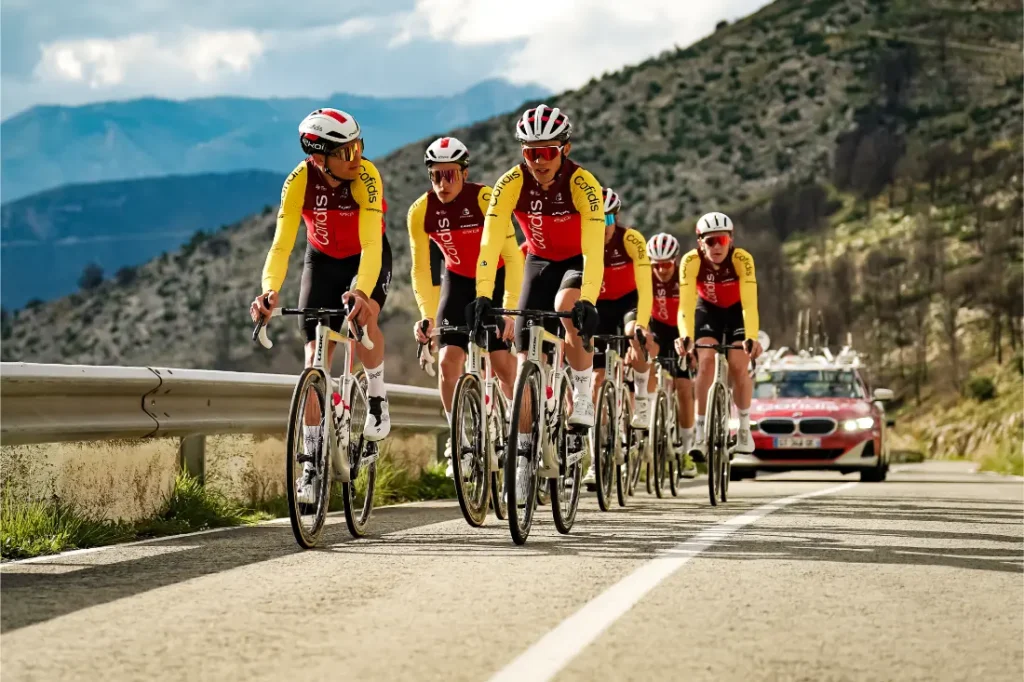 Un grupo de seis ciclistas con uniformes rojos y amarillos a juego, patrocinados por Bicimax, suben por una carretera rural con un coche del equipo Motul Bike Care detrás.