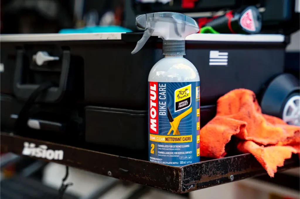 Una botella de Motul Bike Care E9 Wash & Wax Spray reposa sobre una superficie metálica junto a un paño de limpieza naranja, una caja de herramientas negra y una etiqueta de Bicimax.