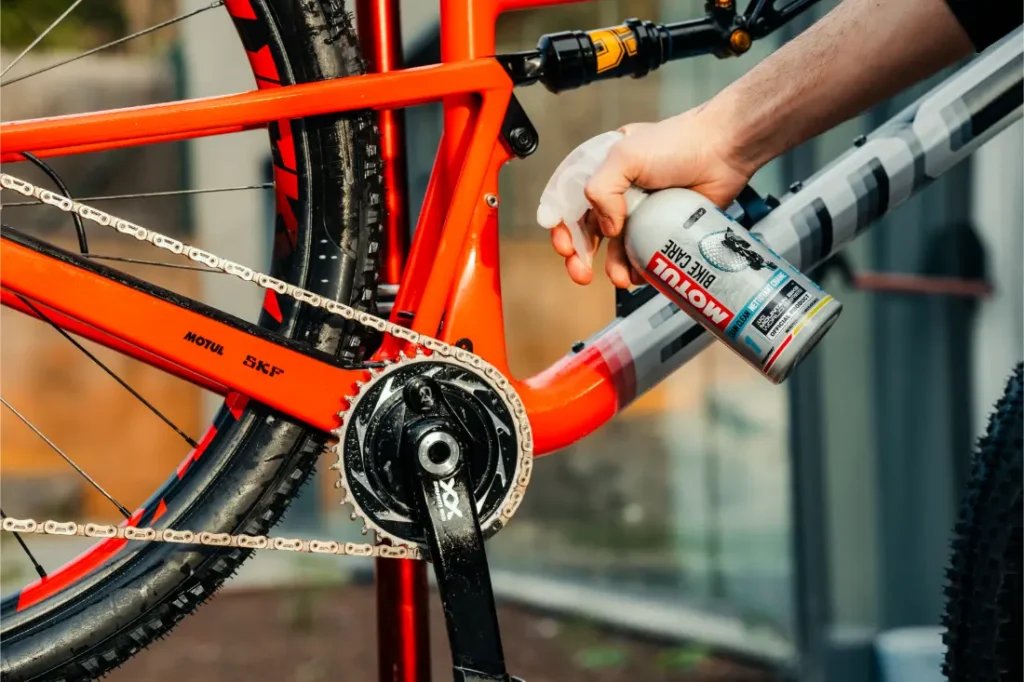 Una persona rocía Motul Bike Care sobre el cuadro de una bicicleta roja y gris, cerca del área de la cigüeñal y la cadena.