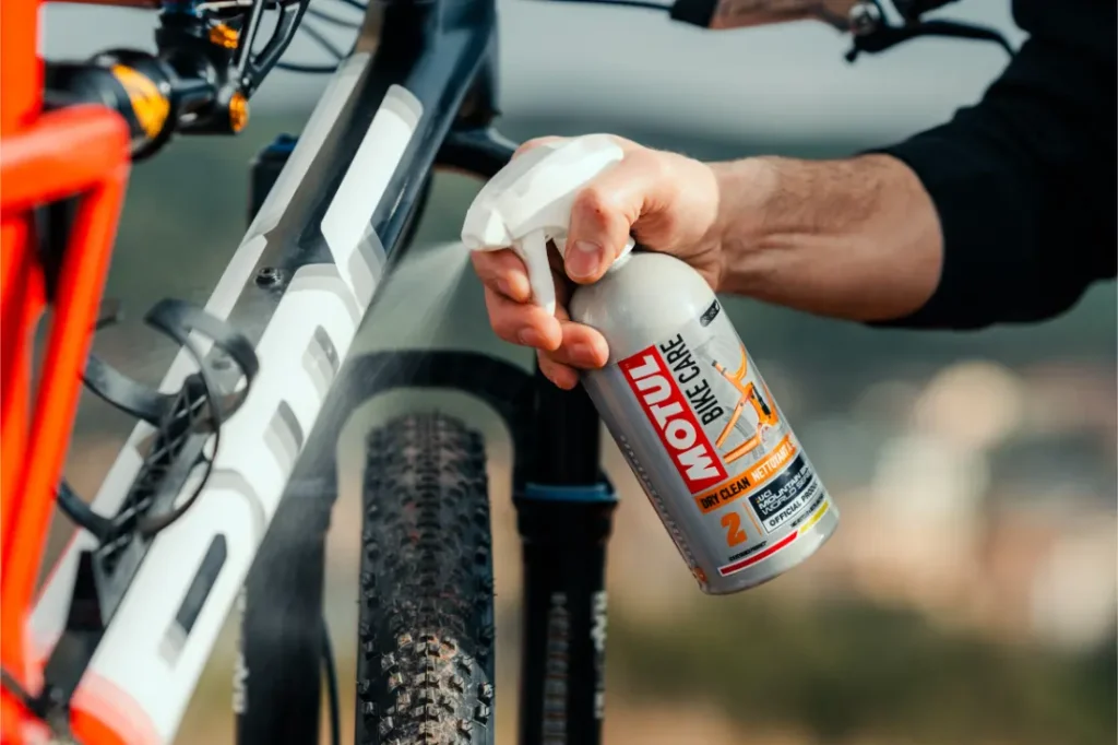 Una persona rocía el limpiador Motul Bike Care sobre el cuadro y el neumático de una bicicleta de montaña al aire libre, manteniendo su bicicleta impecable con el poder de limpieza de Bicimax.