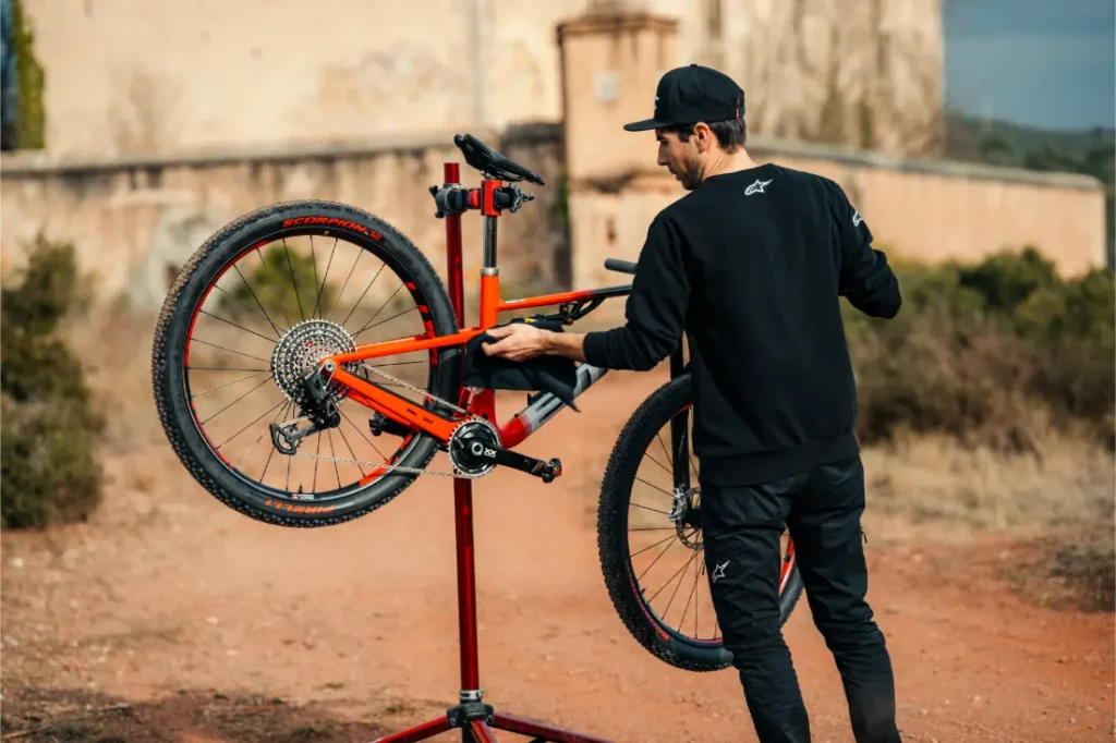 Una persona utiliza productos Motul Bike Care mientras repara o ajusta una bicicleta roja montada en un soporte portátil al aire libre en un camino de tierra.