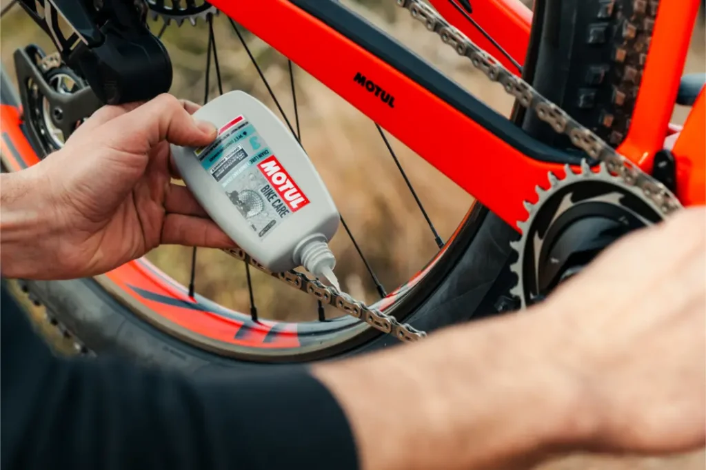 Una persona aplica lubricante Motul Bike Care a la cadena de una bicicleta naranja, centrándose en el mantenimiento de la transmisión con los confiables productos Bicimax.