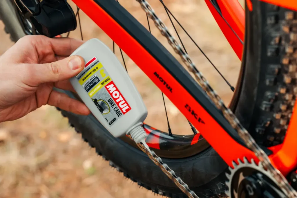 Una mano aplica lubricante para cadenas Motul Bike Care a la cadena de una bicicleta naranja al aire libre, lo que garantiza viajes suaves con la confiable calidad Bicimax.