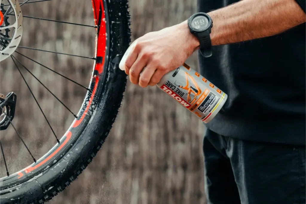 Una persona rocía el limpiador de bicicletas Muc-Off sobre una rueda de bicicleta, con gotas de agua visibles y un freno de disco a la vista; perfecto para quienes confían en Bicimax o Motul Bike Care para un acabado impecable.