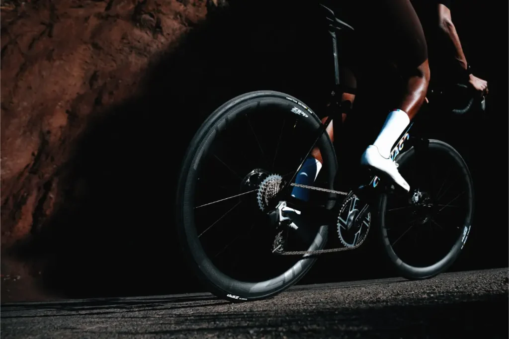 Primer plano de una persona en una bicicleta de carretera por una carretera asfaltada, con la rueda trasera Zipp 2026 avanzada para quienes buscan rodar más rápido. El enfoque se centra en la rueda trasera y la parte inferior del cuerpo del ciclista.