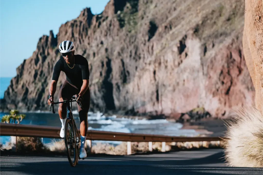 Un ciclista con casco y ropa de ciclismo negra conduce una bicicleta de ruta Zipp 2026 por una carretera costera con acantilados y el océano de fondo, buscando rodar más rápido en esta ruta escénica.