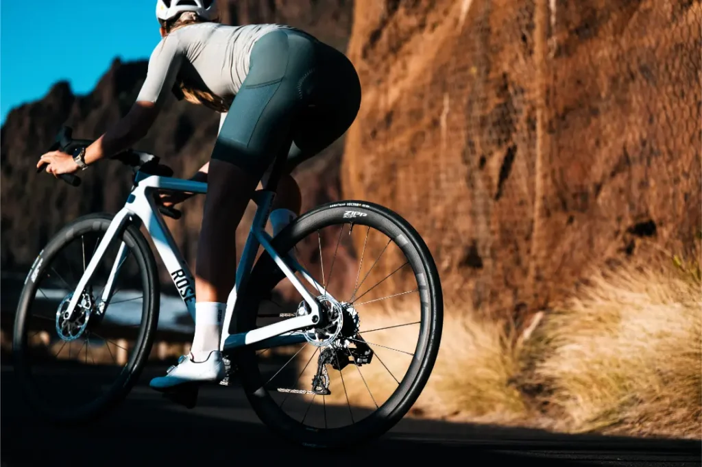 Un ciclista con casco y equipo de ciclismo circula en una bicicleta de carretera blanca equipada con ruedas Zipp 2026 por una carretera asfaltada junto a un acantilado rocoso e iluminado por el sol, dispuesto a rodar más rápido en cada tramo.