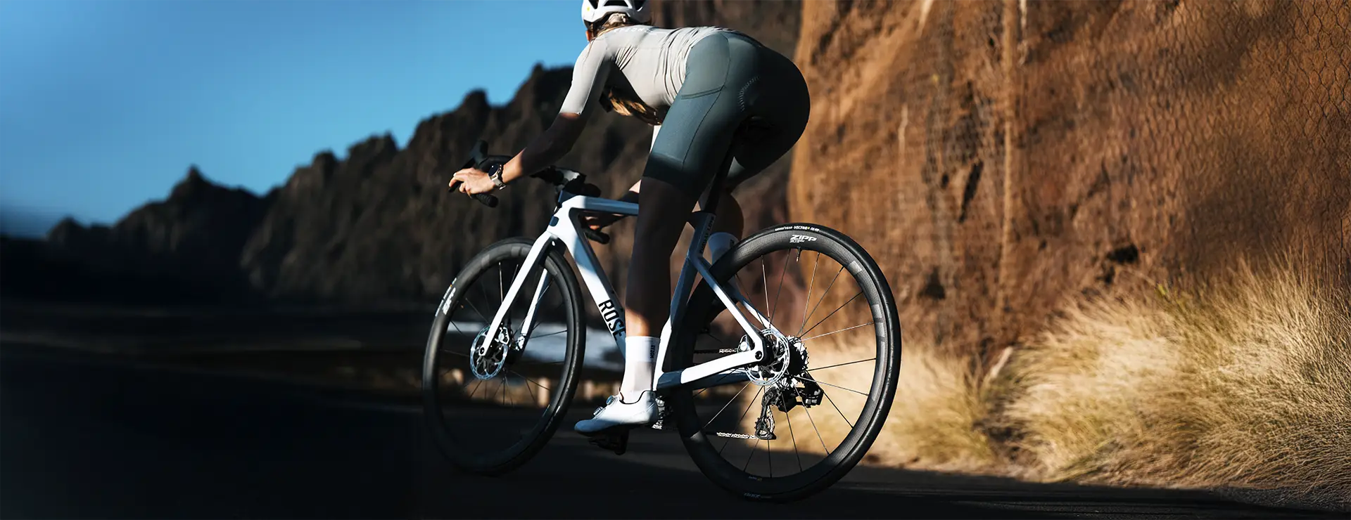 Un ciclista circula en una bicicleta de carretera blanca con ruedas Zipp 2026 por una carretera asfaltada junto a acantilados rocosos y hierba seca, usando casco y equipo de ciclismo para rodar más rápido.