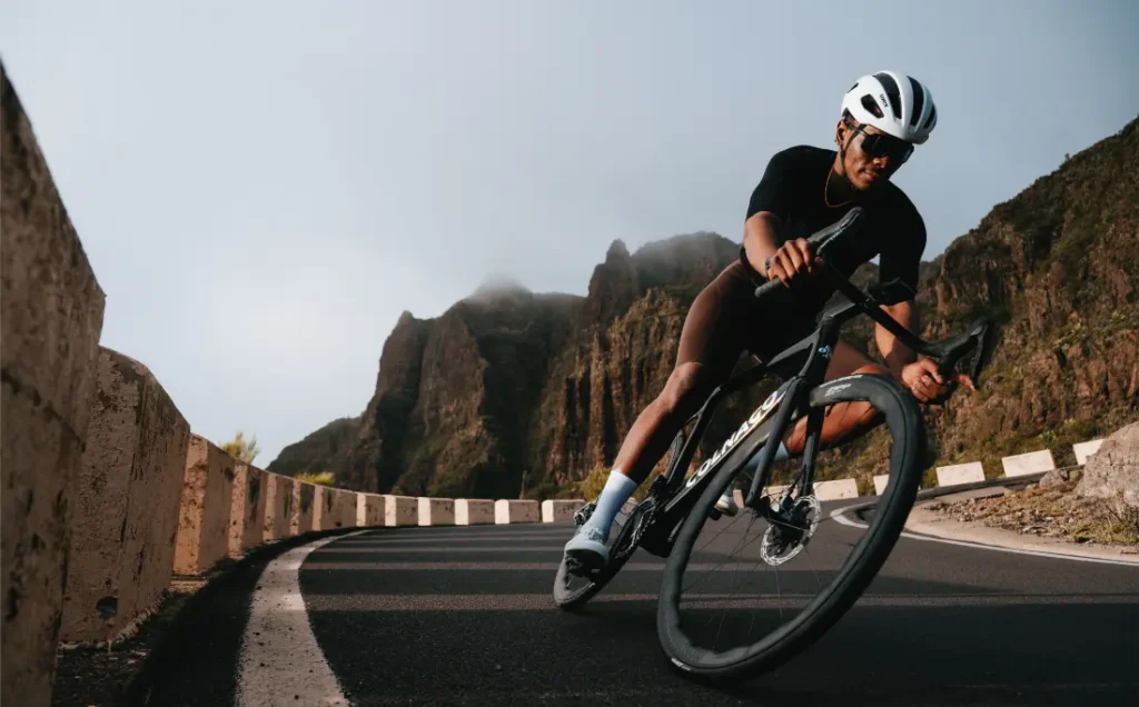 Un ciclista con casco y gafas de sol conduce una bicicleta de carretera Zipp 2026 por una curva en una carretera de montaña con acantilados rocosos al fondo, listo para rodar más rápido.