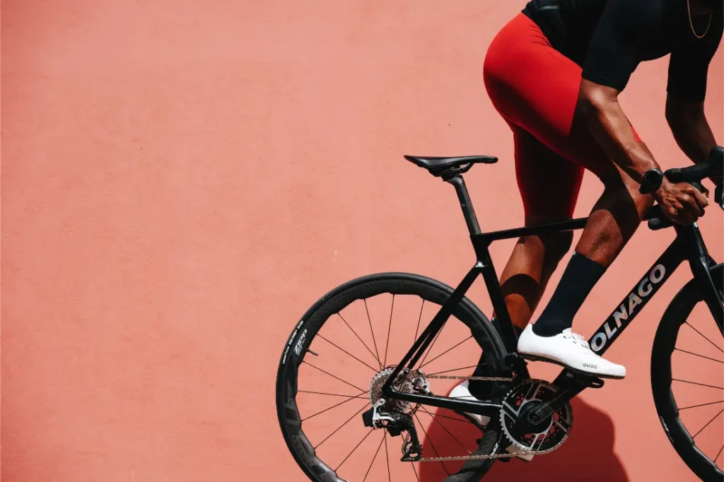 Una persona que viste pantalones cortos de ciclismo de color rojo monta una bicicleta de carretera Colnago negra con ruedas Zipp 2026, lista para rodar más rápido contra una pared sólida de color melocotón.
