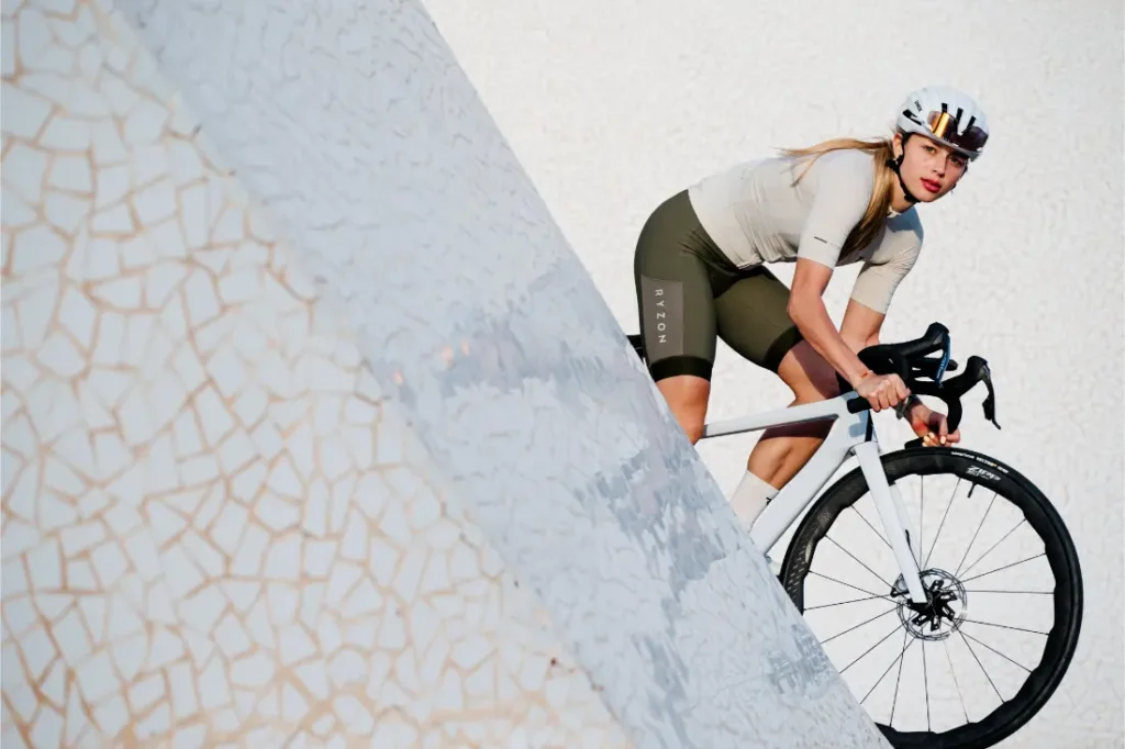 Un ciclista con casco y equipo de ciclismo conduce una bicicleta blanca equipada con ruedas Zipp 2026 junto a una estructura grande, alicatada y angular con un fondo neutro, listo para rodar más rápido.