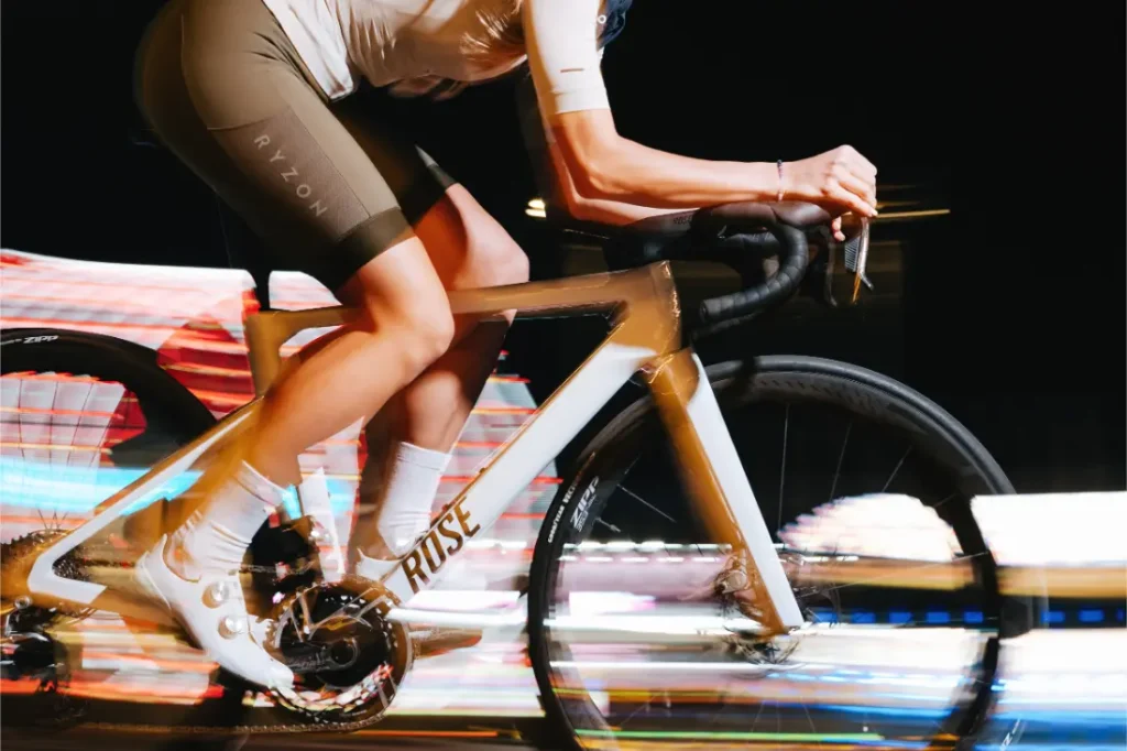 Un ciclista con pantalones cortos marrones y maillot blanco avanza a toda velocidad en la noche sobre una bicicleta Rose blanca con ruedas Zipp 2026, luces borrosas que insinúan rodar más rápido y un rendimiento inigualable.