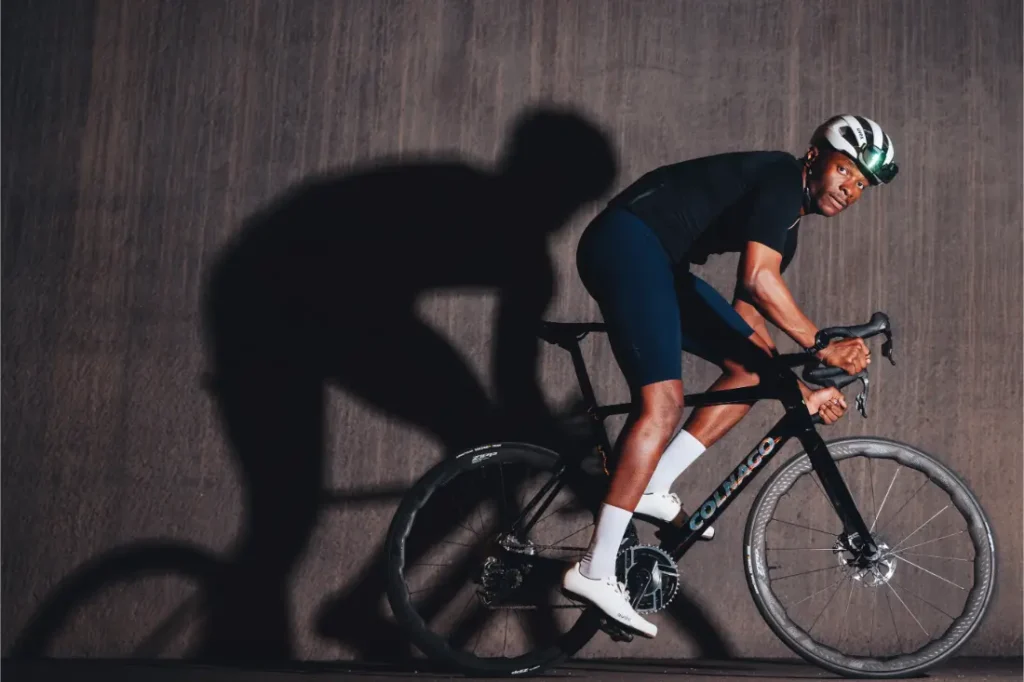 Un ciclista vestido con traje y casco negros conduce una bicicleta de ruta Zipp 2026 contra un fondo oscuro y liso, proyectando una sombra prominente en la pared, listo para rodar más rápido.