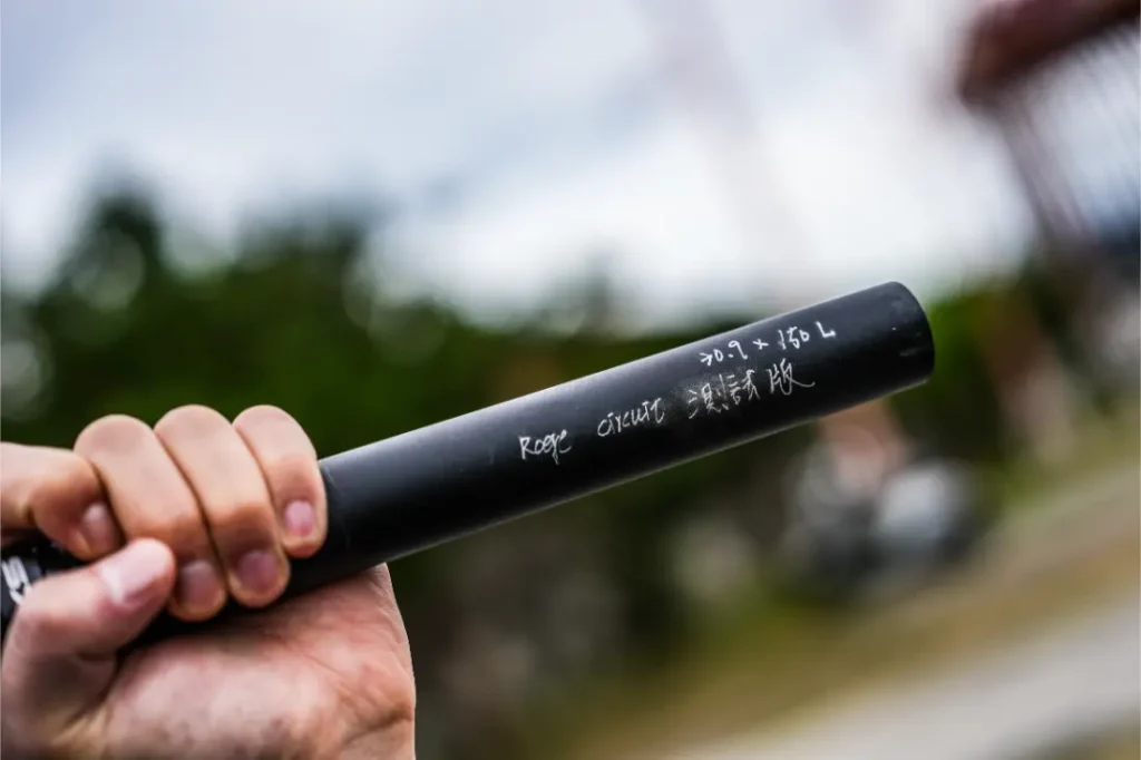 Una mano sostiene una tija telescópica inalámbrica negra RAGE Circuit con letras blancas en inglés y caracteres japoneses sobre un fondo borroso al aire libre.