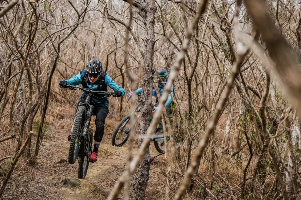 Dos ciclistas de montaña recorren un sendero forestal, con el ciclista que va en cabeza levantando la rueda delantera del suelo gracias a una tija telescópica inalámbrica RAGE Circuit y el segundo ciclista siguiéndole de cerca.